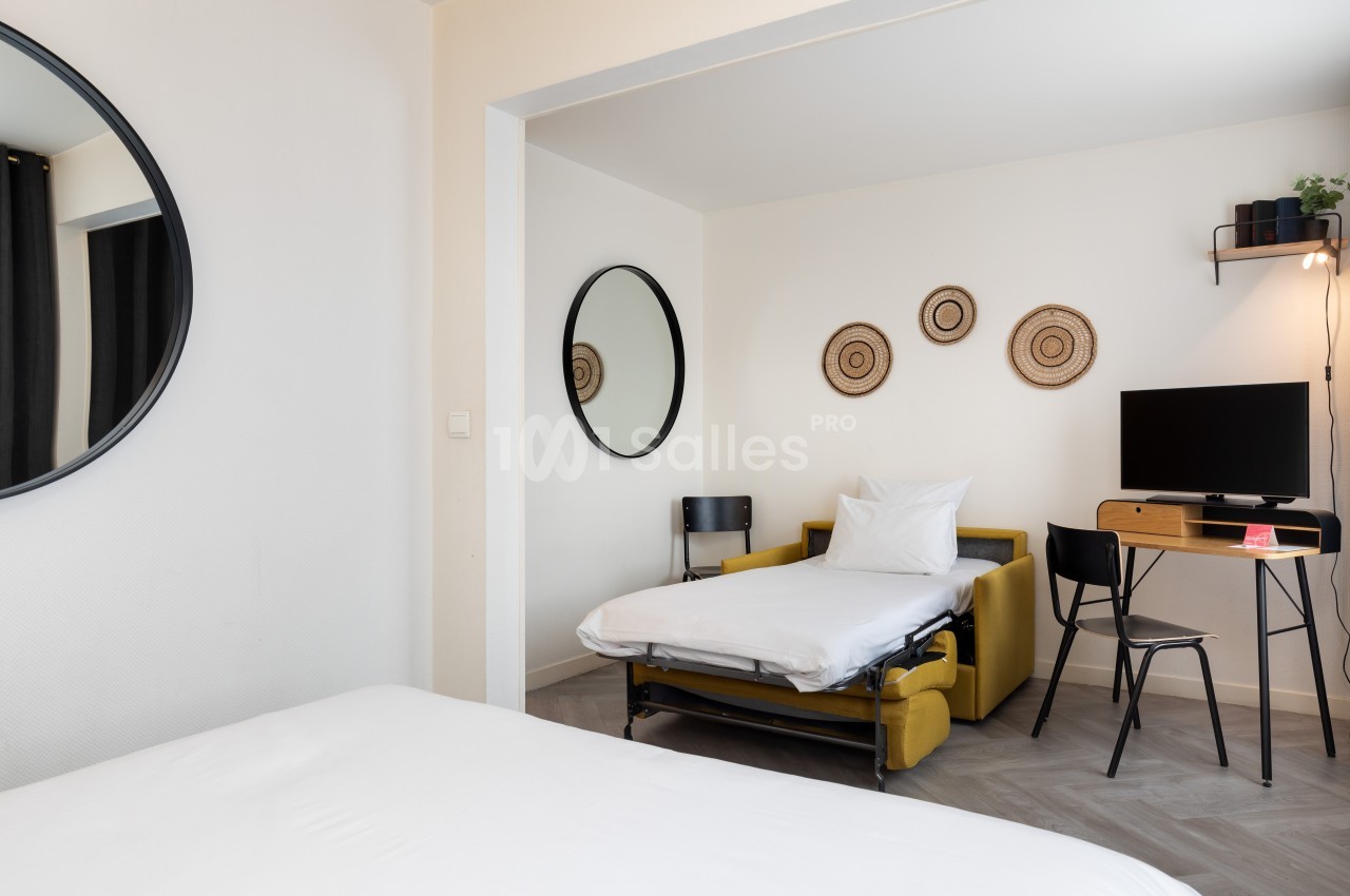 Chambre lumineuse avec un lit déplié, un bureau avec écran, des miroirs ronds et des décorations murales en osier.