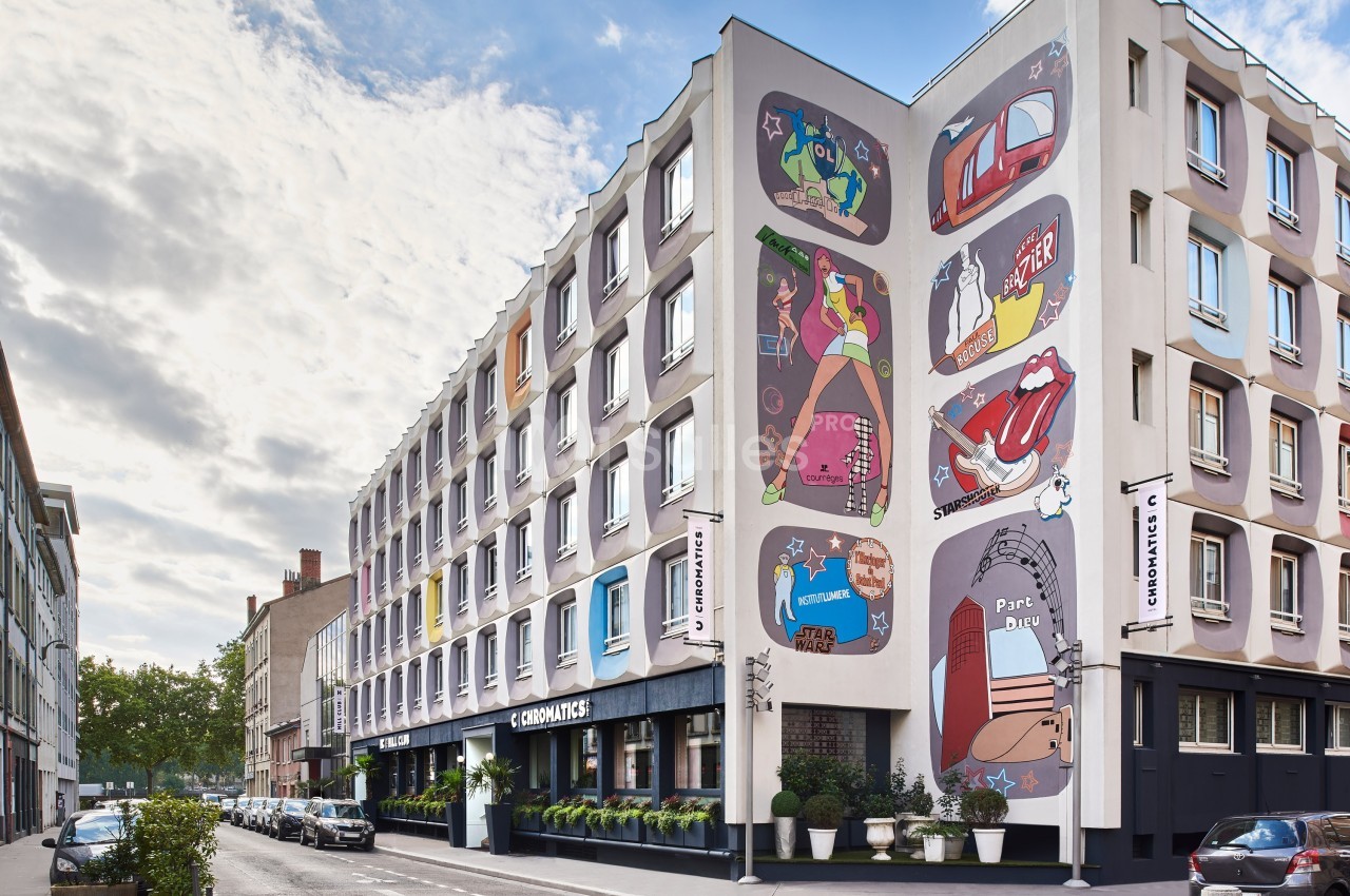 Façade d'un immeuble urbain décorée de fresques colorées, avec une rue bordée de voitures et de végétation.