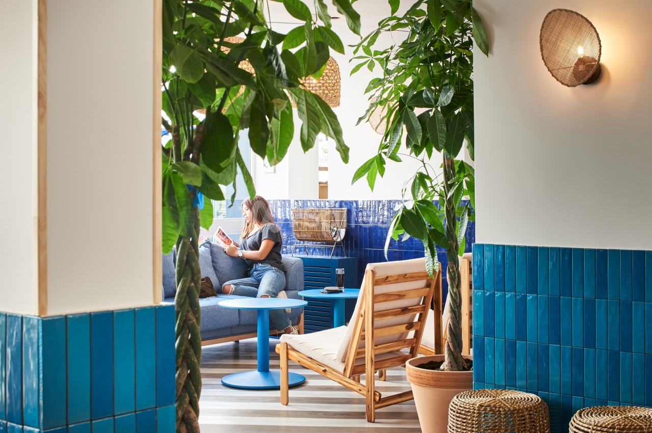 Espace intérieur lumineux avec fauteuils en bois, tables bleues, plantes vertes et une personne lisant sur un canapé.