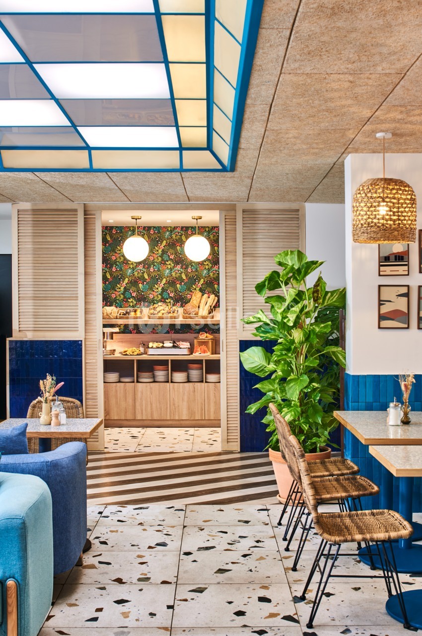 Espace intérieur lumineux avec mobilier en rotin, plantes vertes et vue sur un buffet décoré d'un papier peint floral.