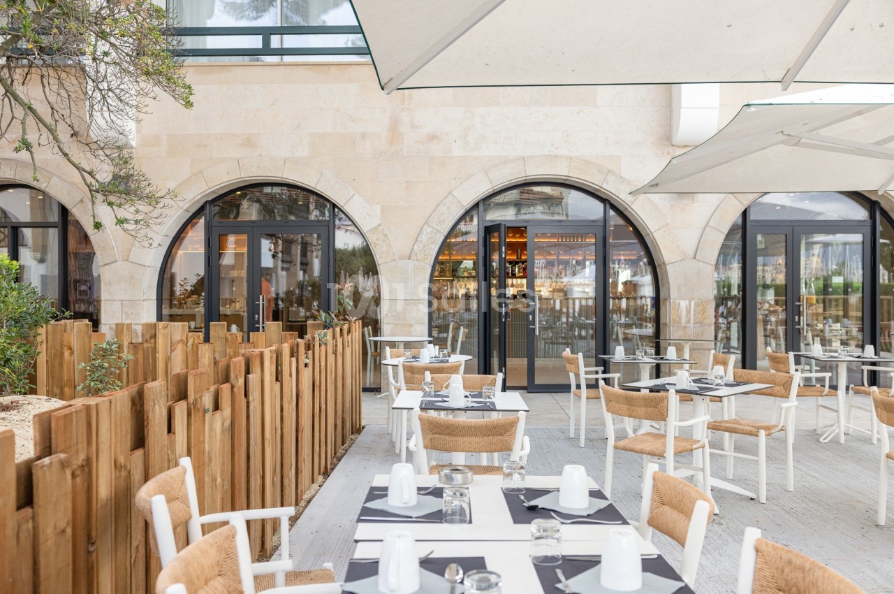 Terrasse d'un restaurant avec tables dressées, chaises en bois et parasols, devant une façade en pierre avec grandes baies…