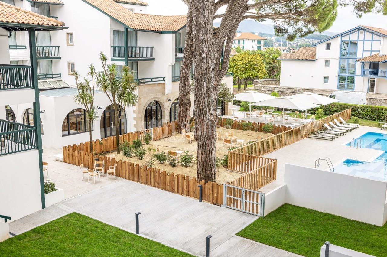 Cour intérieure d'une résidence avec terrasse, piscine, espaces verts, et clôture en bois autour d'une aire de jeux.