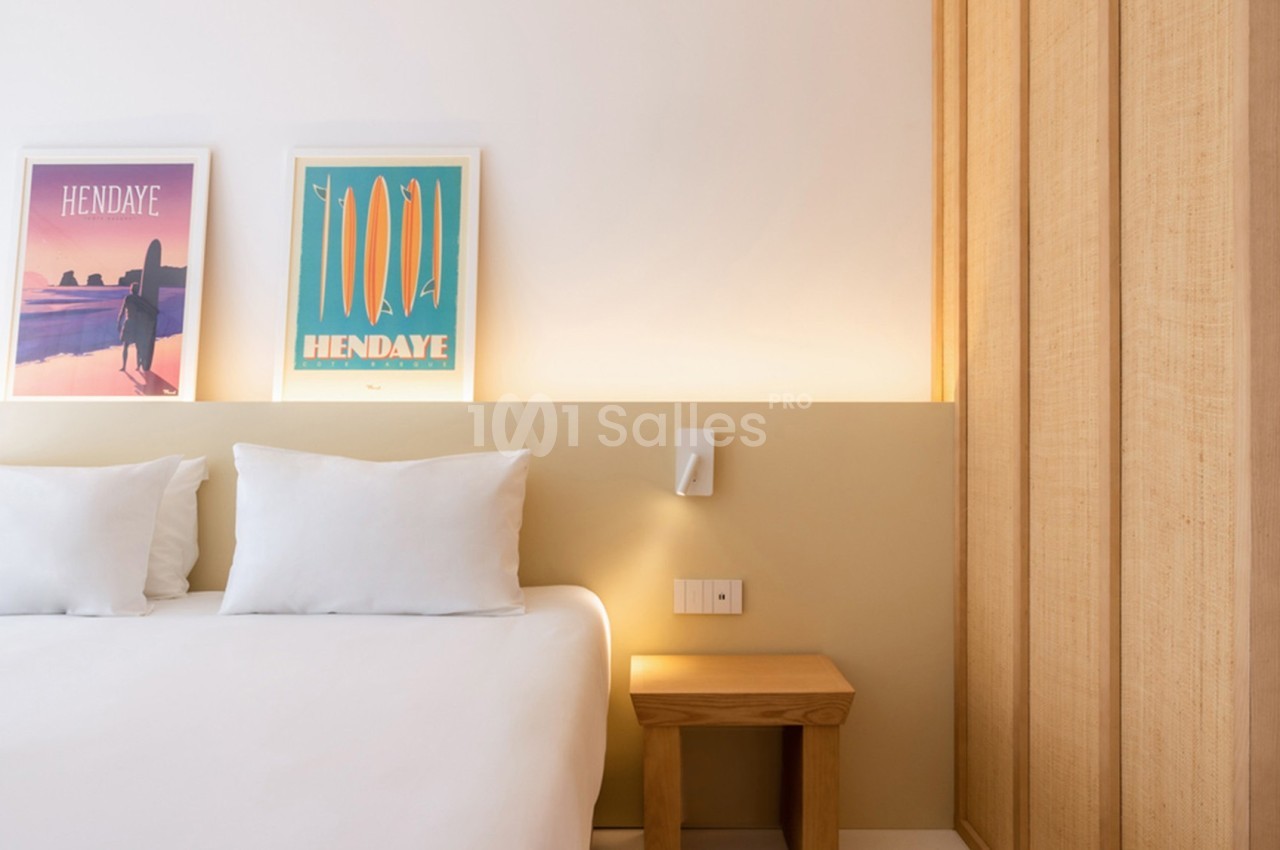 Lit double avec draps blancs, table de chevet en bois et affiches encadrées sur un mur clair dans une chambre lumineuse.