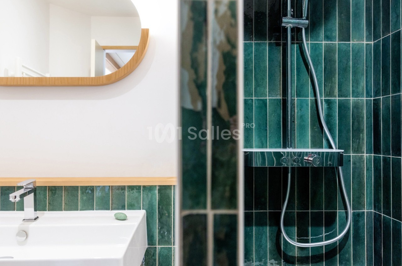 Lavabo blanc avec miroir en bois et douche à carreaux verts dans une salle de bain moderne.