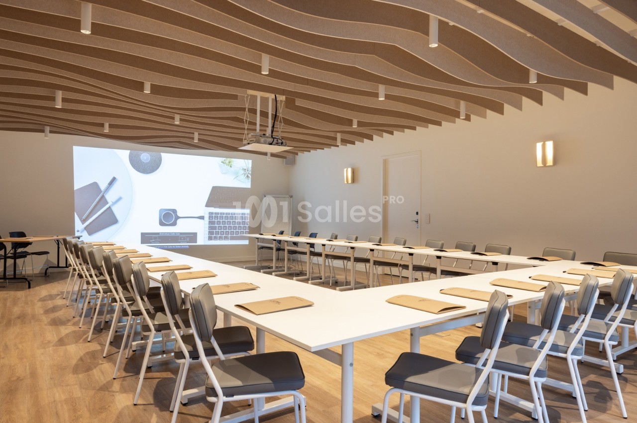 Salle de réunion moderne avec tables disposées en U, chaises alignées et projection d'une présentation sur un écran.