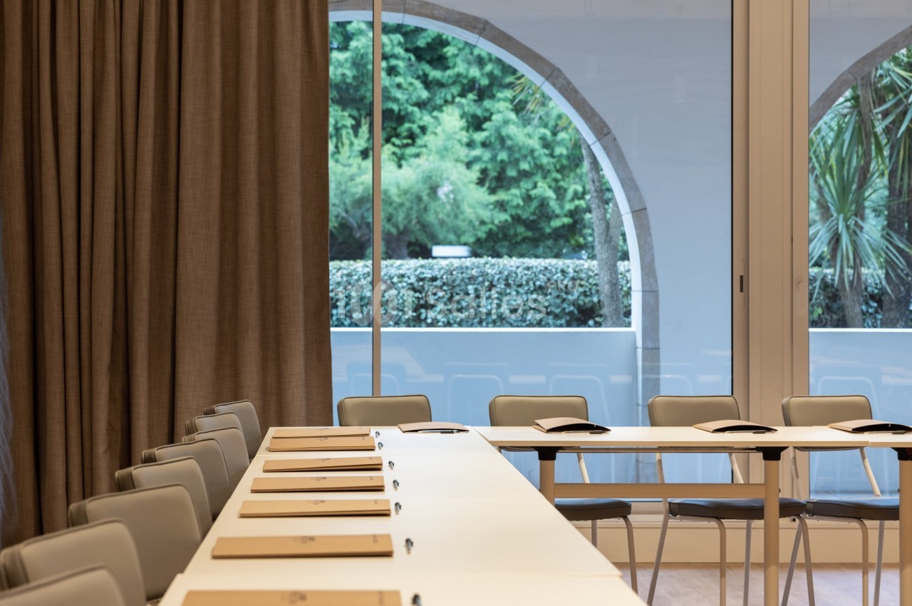 Salle de réunion lumineuse avec des tables blanches alignées, des chaises grises et une grande baie vitrée donnant sur un…
