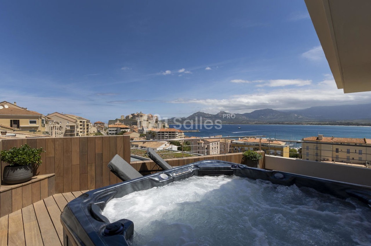 Vue d'une terrasse avec jacuzzi, surplombant une ville côtière, la mer et des montagnes sous un ciel dégagé.