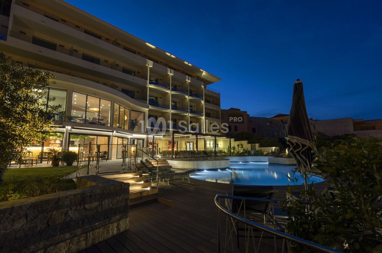 Hôtel moderne éclairé de nuit avec piscine extérieure et terrasse en bois entourée de végétation.