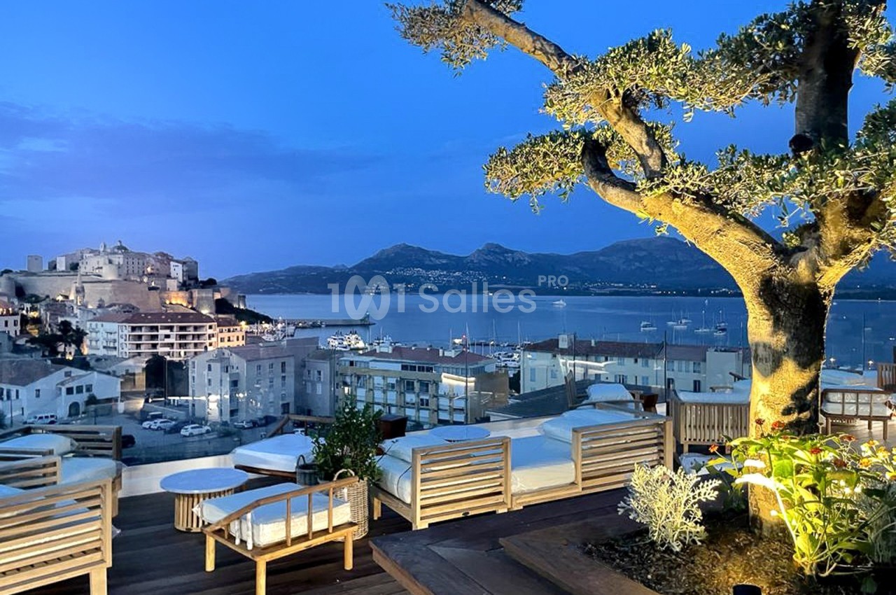 Vue sur une terrasse aménagée avec des fauteuils en bois, donnant sur un port, une ville et des montagnes au crépuscule.