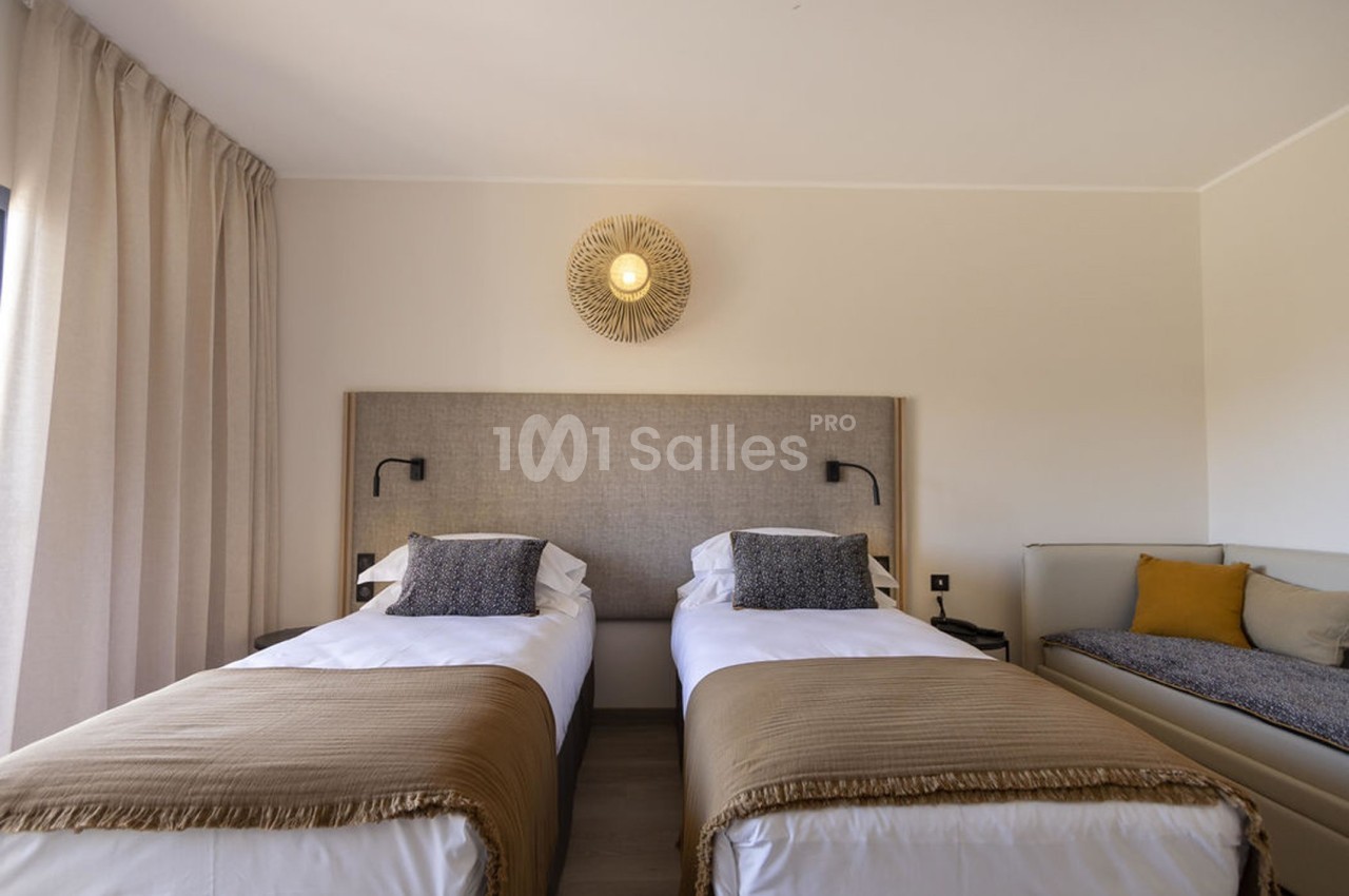 Deux lits simples avec des couvre-lits beige, une applique murale et un canapé avec coussins dans une chambre lumineuse.