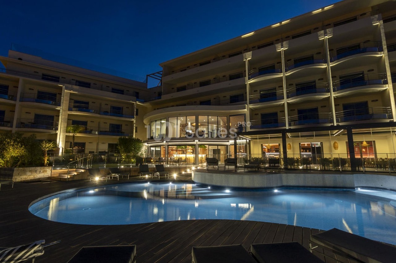 Façade d'un hôtel éclairé de nuit avec une piscine au premier plan entourée de transats.