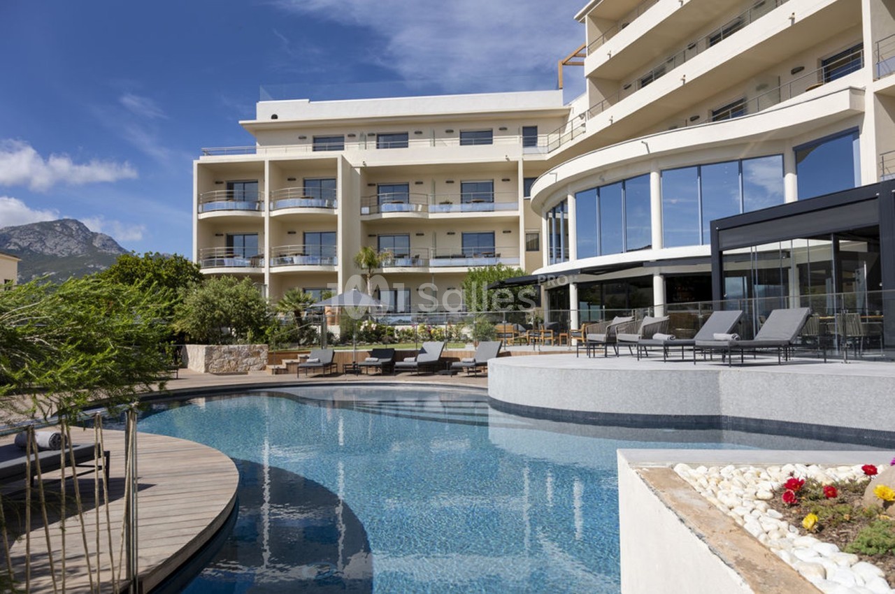 Piscine extérieure bordée de chaises longues, devant un bâtiment moderne avec balcons, sous un ciel bleu.