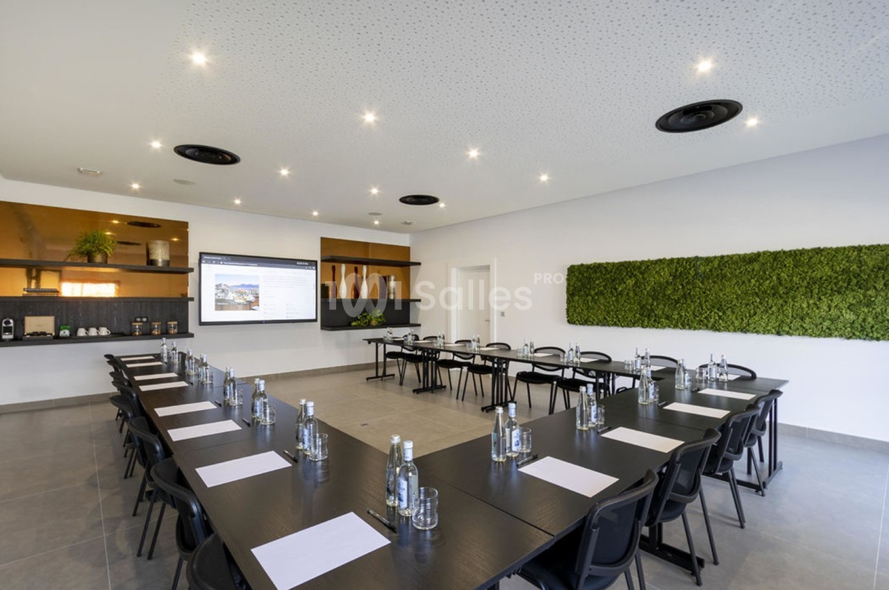Salle de réunion moderne avec tables en U, bouteilles d'eau, blocs-notes, écran de présentation et mur végétal.