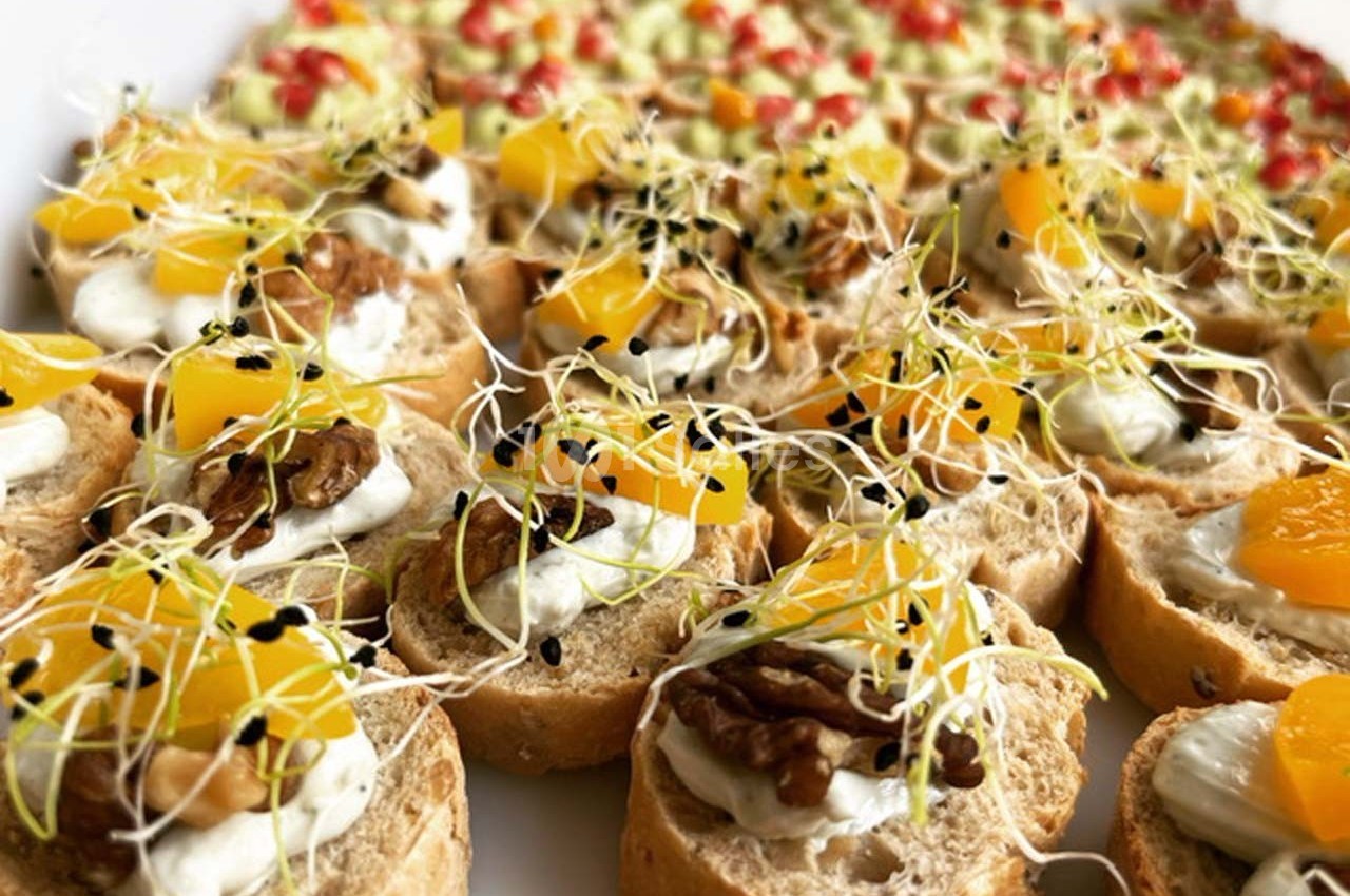 Assortiment de canapés garnis de fromage frais, noix, morceaux de fruits et pousses, disposés sur un plateau.