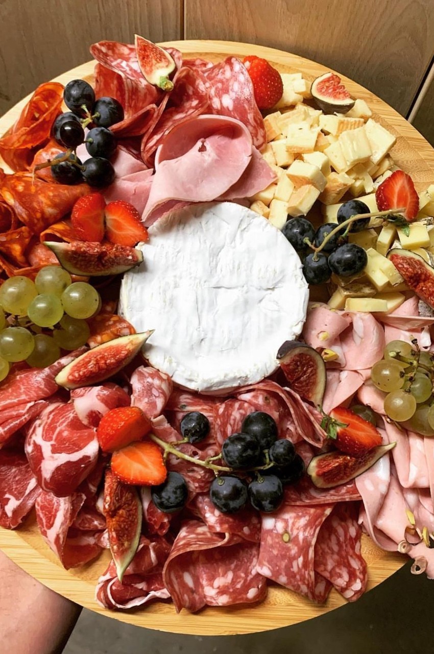 Plateau rond avec charcuteries variées, fromages, raisins, fraises et figues autour d’un fromage à pâte molle au centre.