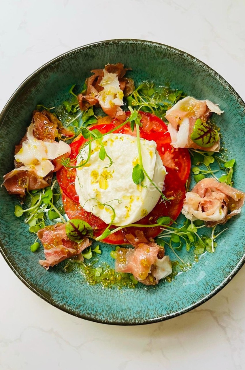 Assiette garnie de burrata, tranches de tomate, jambon cru, herbes fraîches et filet d'huile sur un fond clair.