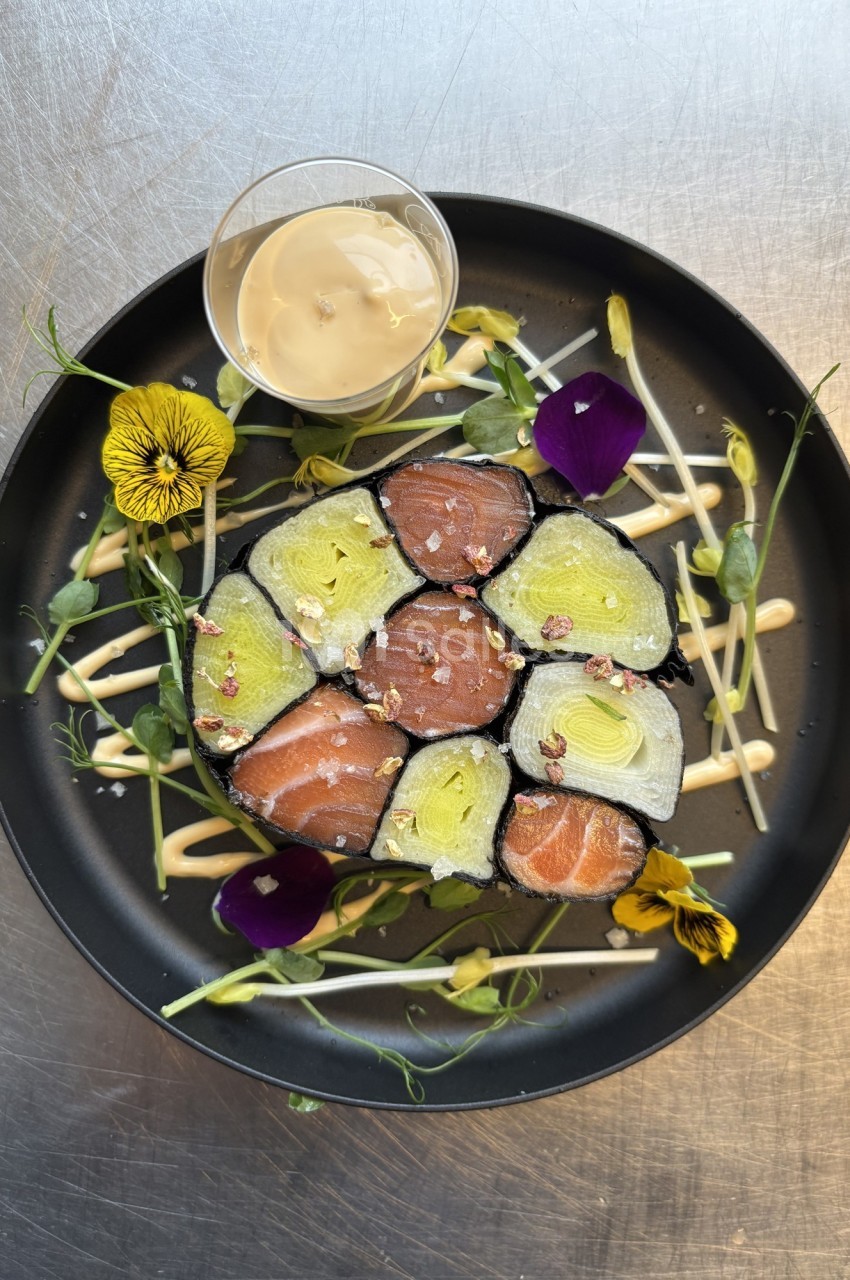 Assiette de makis colorés garnis de saumon et légumes, décorée de fleurs comestibles et accompagnée d'une sauce.