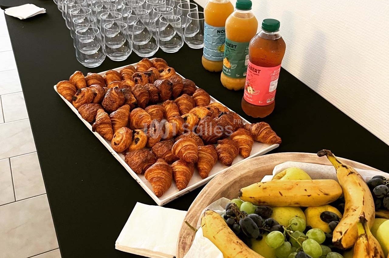 Plateau de viennoiseries, corbeille de fruits, bouteilles de jus et verres vides disposés sur une table noire.