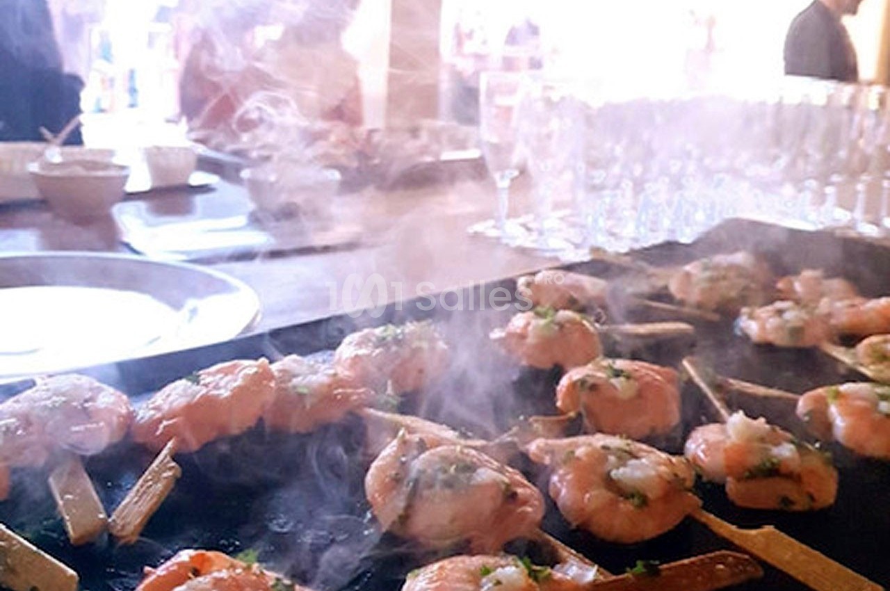 Brochettes de saumon et crevettes grillées sur une plancha, entourées de vapeur, dans un cadre de réception.