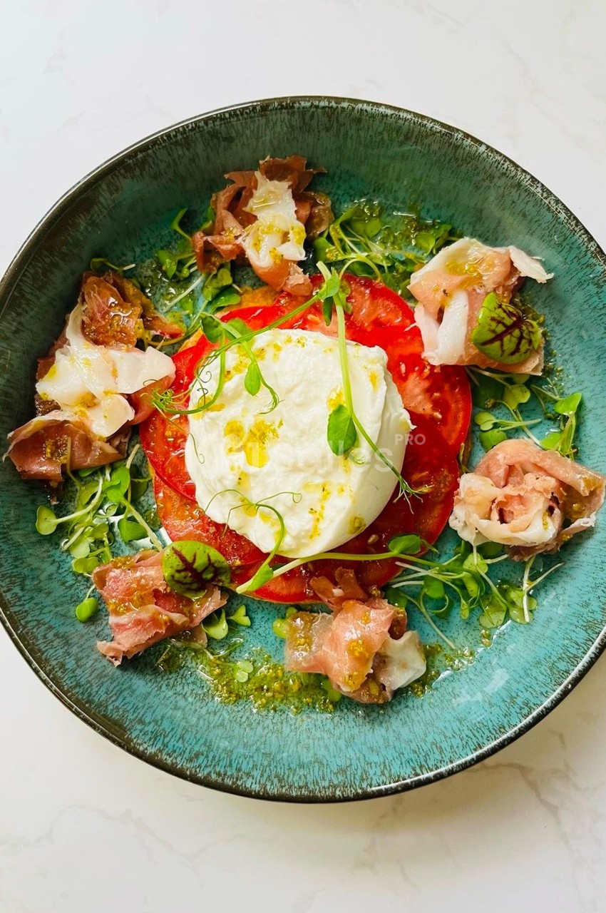 Assiette garnie de burrata, tranches de tomate, jambon cru, herbes fraîches et filet d'huile sur un fond clair.