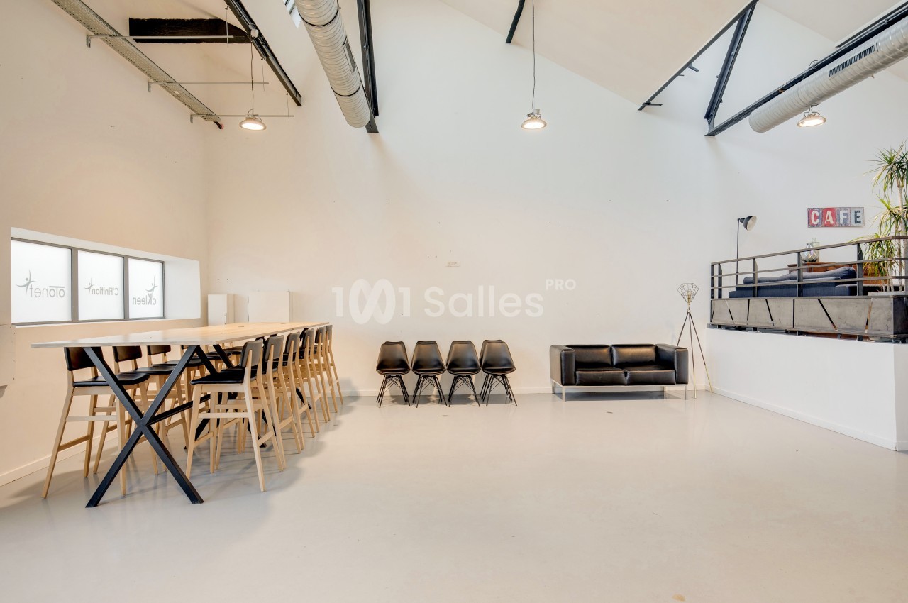 Salle lumineuse avec de grandes fenêtres, table en bois entourée de chaises, canapé noir et mezzanine avec décoration.