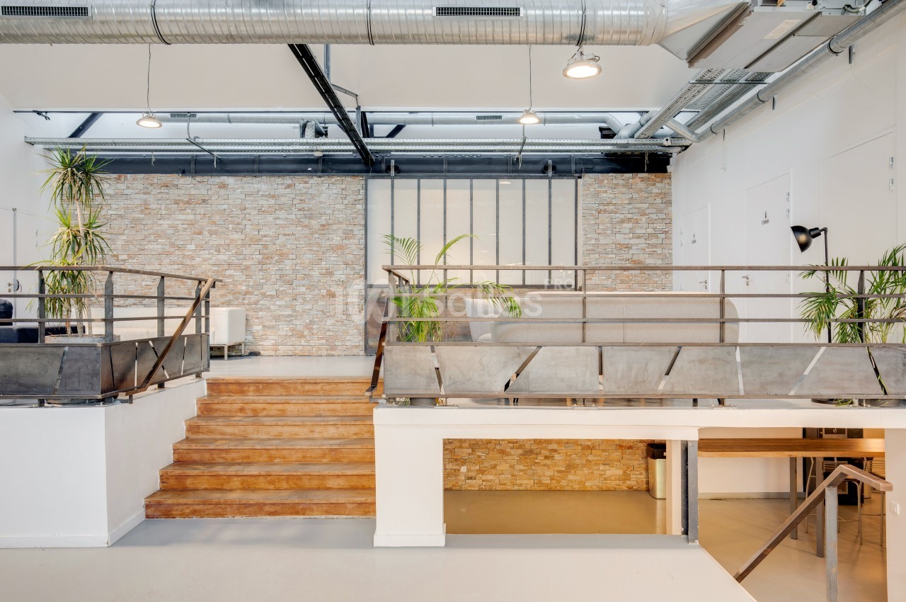 Espace intérieur lumineux avec murs en briques, mezzanine en métal, plantes et escalier en bois.