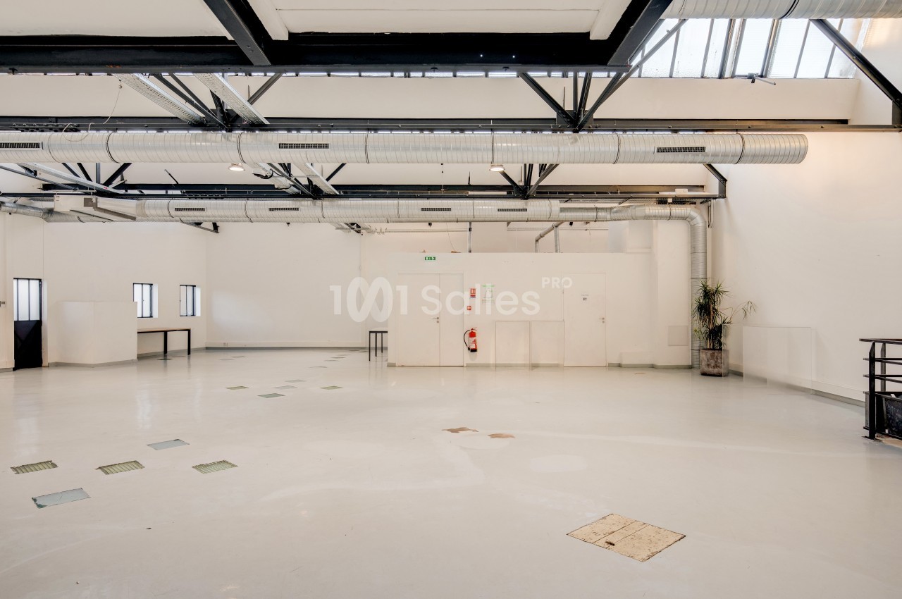Intérieur d'un grand espace industriel lumineux avec sol clair, murs blancs, poutres métalliques et conduits apparents.