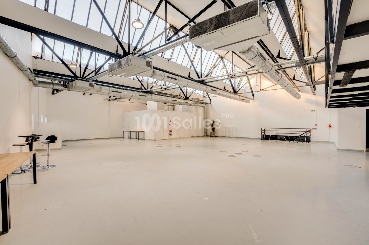 Grand espace intérieur lumineux avec plafond en verrière, poutres métalliques apparentes et sol en béton clair.