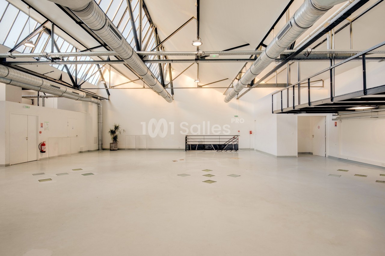 Grand espace intérieur lumineux avec plafond haut, conduits apparents, murs blancs et mezzanine métallique.