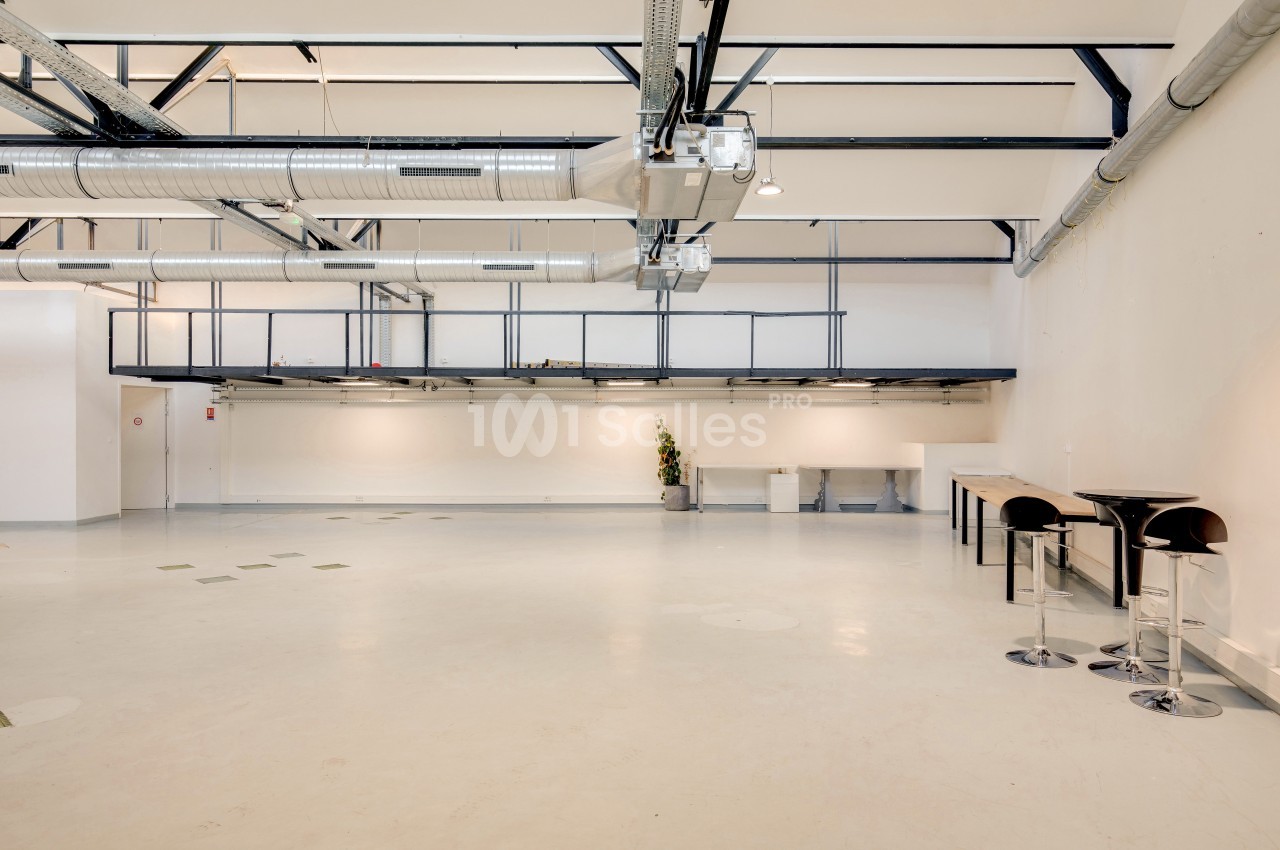 Intérieur d'un espace industriel lumineux avec plafond haut, conduits apparents et mobilier minimaliste.