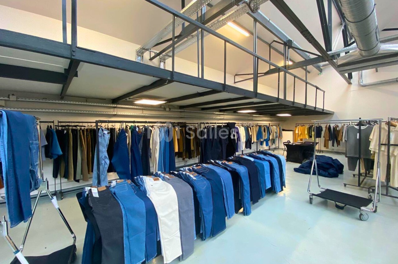 Portants de vêtements variés, principalement des jeans, exposés dans un espace lumineux et industriel.