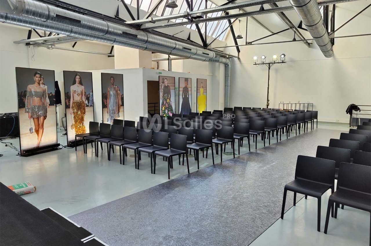 Salle de défilé vide avec rangées de chaises noires, podium central et affiches de mannequins en arrière-plan.