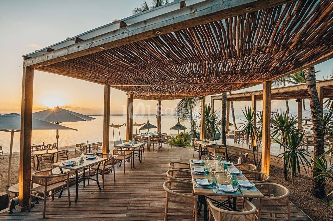 Terrasse en bois d'un restaurant en bord de mer, avec des tables dressées et vue sur le coucher de soleil.