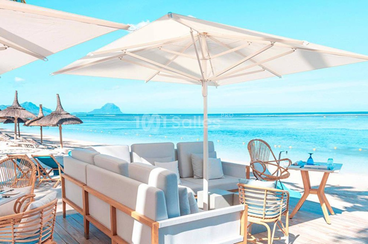Terrasse en bord de mer avec parasols, mobilier en bois et vue sur une plage de sable et une eau turquoise.