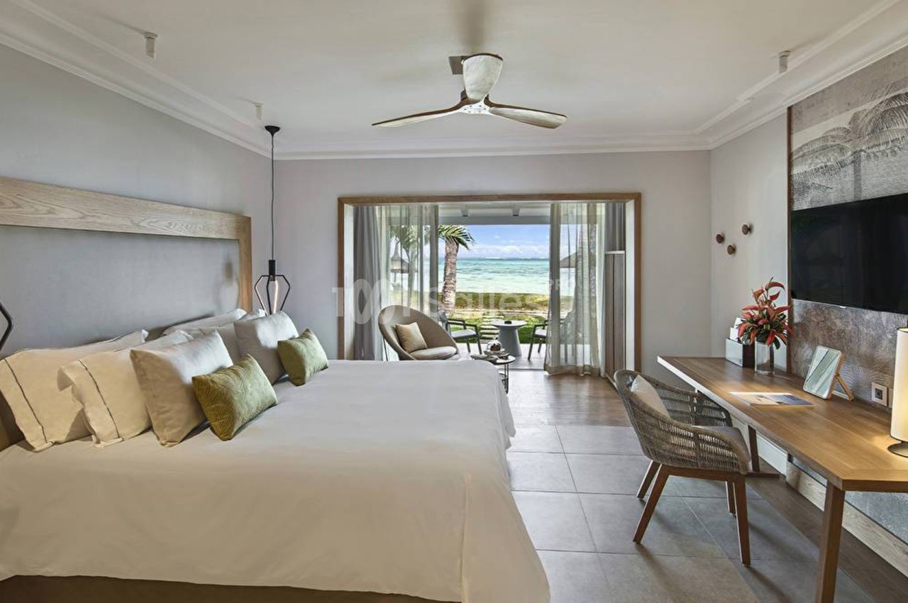 Chambre lumineuse avec lit double, bureau, fauteuils et vue sur la mer à travers une baie vitrée.