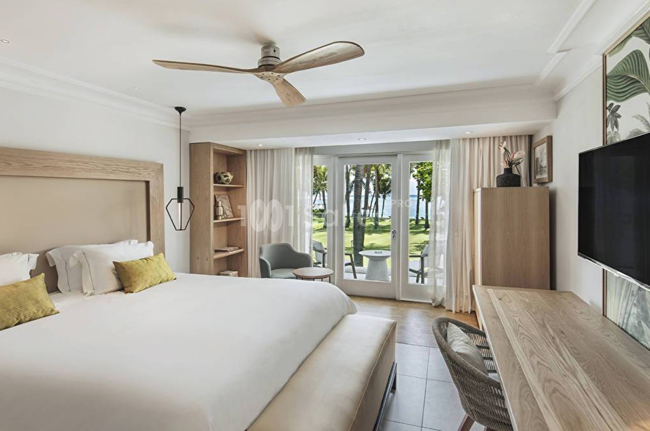 Chambre lumineuse avec lit double, bureau, télévision, ventilateur au plafond et vue sur un jardin menant à la mer.