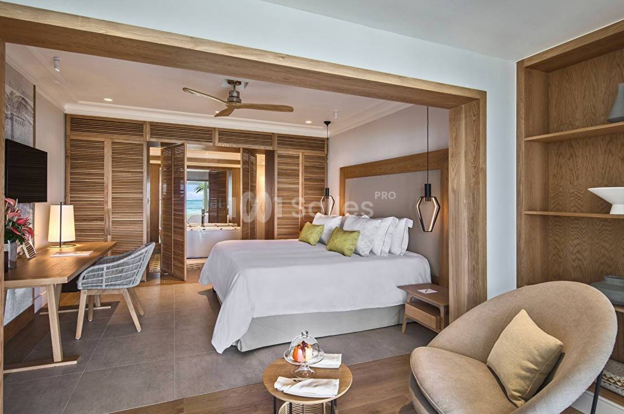 Chambre lumineuse avec lit double, bureau, fauteuil, étagères en bois et vue sur une salle de bain ouverte.