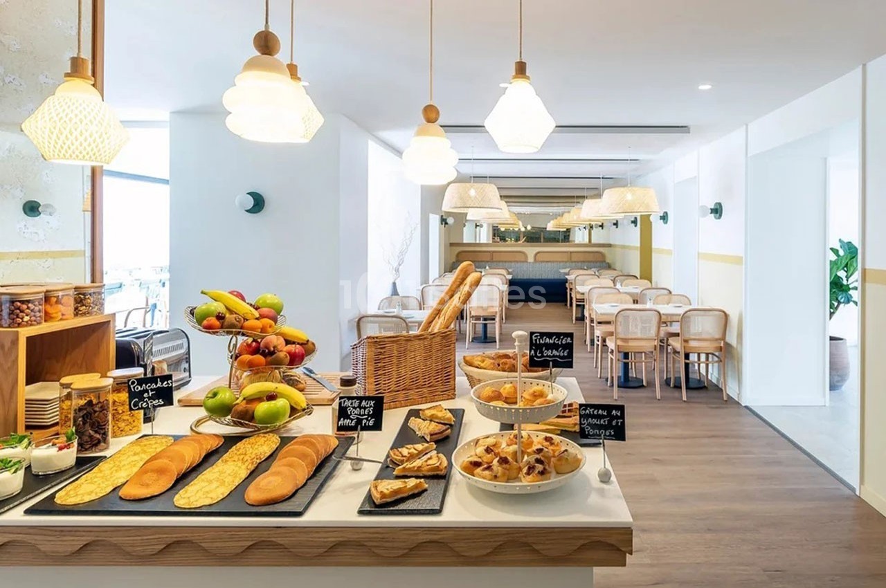 Buffet de petit-déjeuner avec viennoiseries, fruits, pains et céréales dans une salle lumineuse et moderne.