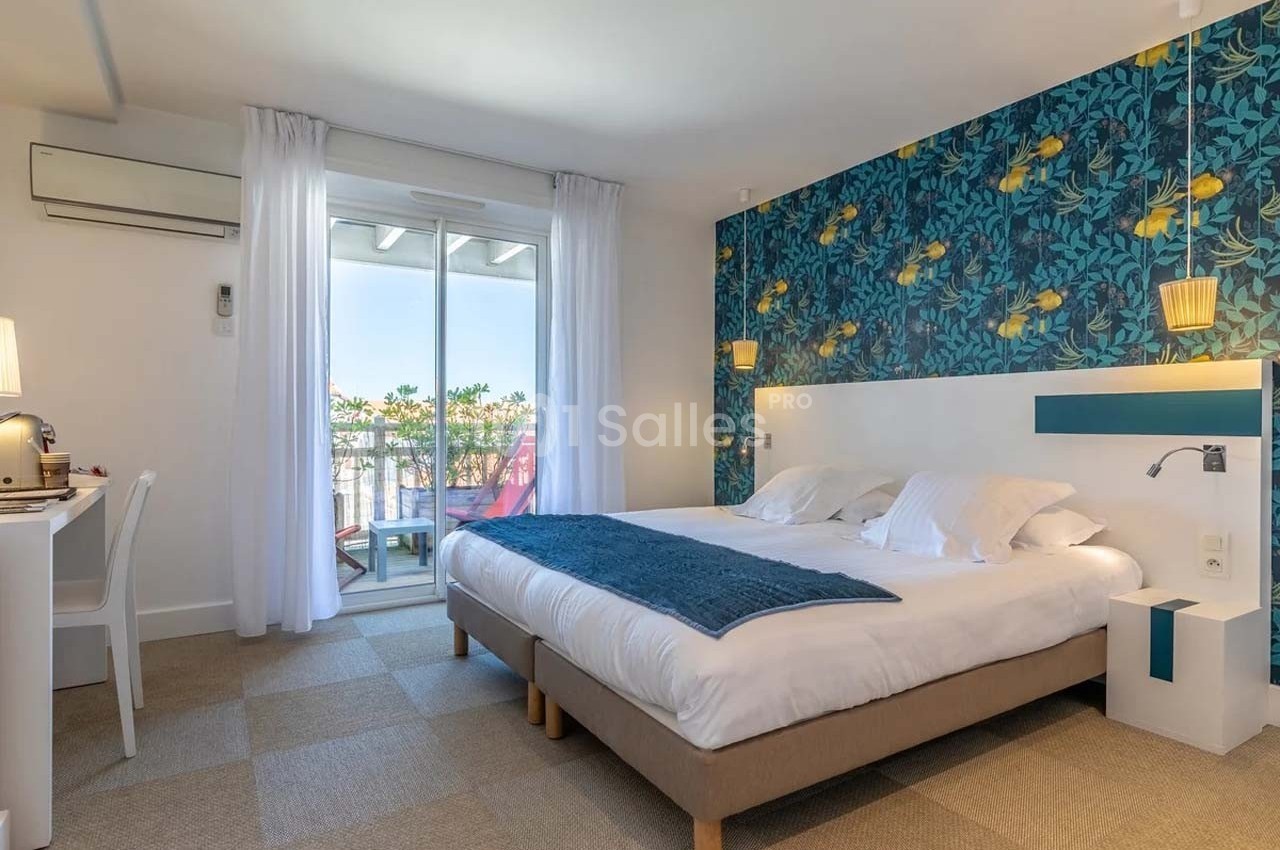 Chambre lumineuse avec lit double, bureau, mur décoré de motifs floraux et accès à un balcon avec vue extérieure.