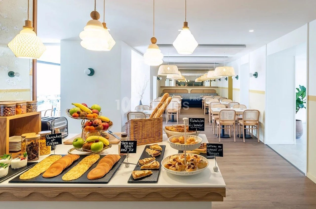 Buffet de petit-déjeuner avec viennoiseries, fruits, pains et céréales dans une salle lumineuse et moderne.