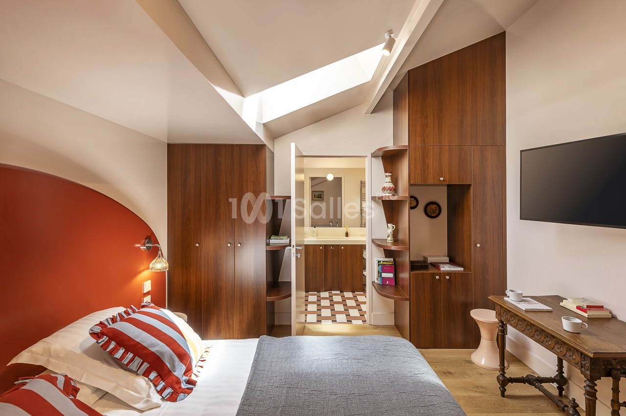 Chambre lumineuse avec lit double, mobilier en bois, mur rouge, télévision murale et salle de bain attenante.