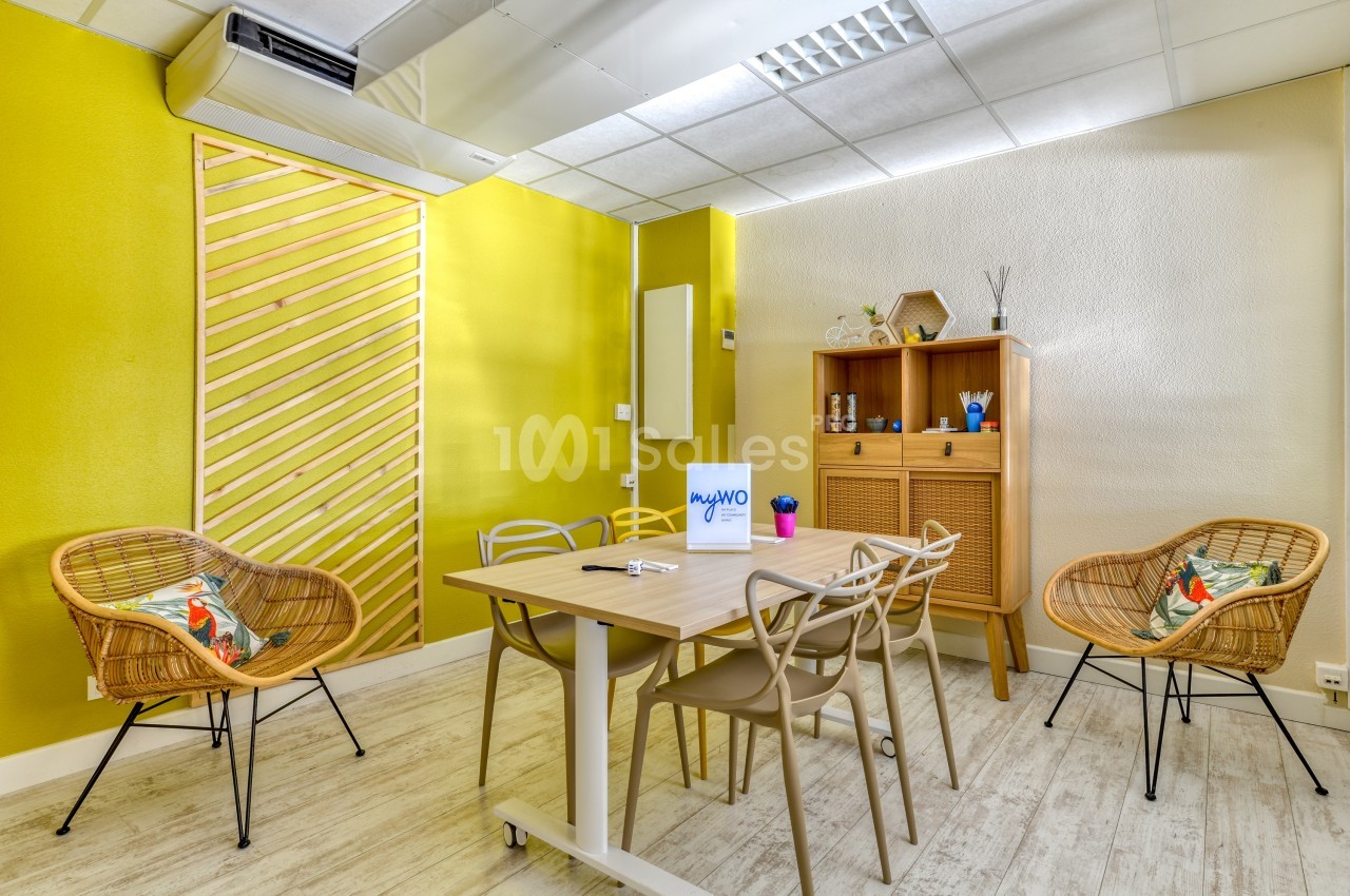 Salle de réunion lumineuse avec table en bois, chaises variées, murs jaunes et mobilier en rotin.