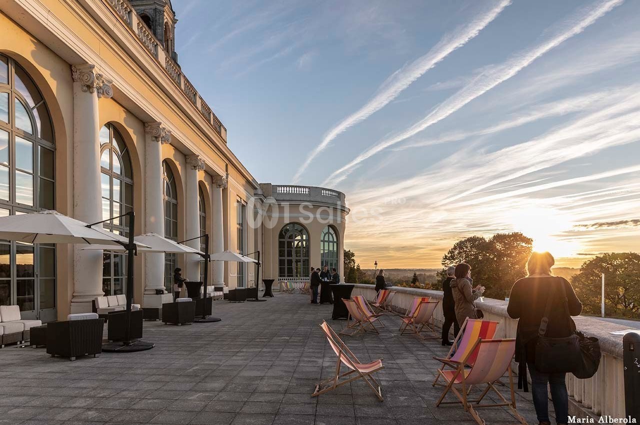 Terrasse avec chaises longues, parasols et vue sur un coucher de soleil, près d'un bâtiment aux grandes baies vitrées.