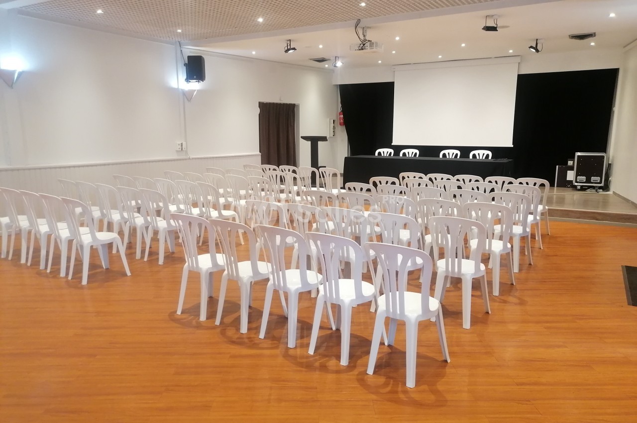 Salle de conférence avec des rangées de chaises blanches en plastique, un podium et un écran de projection à l'arrière.