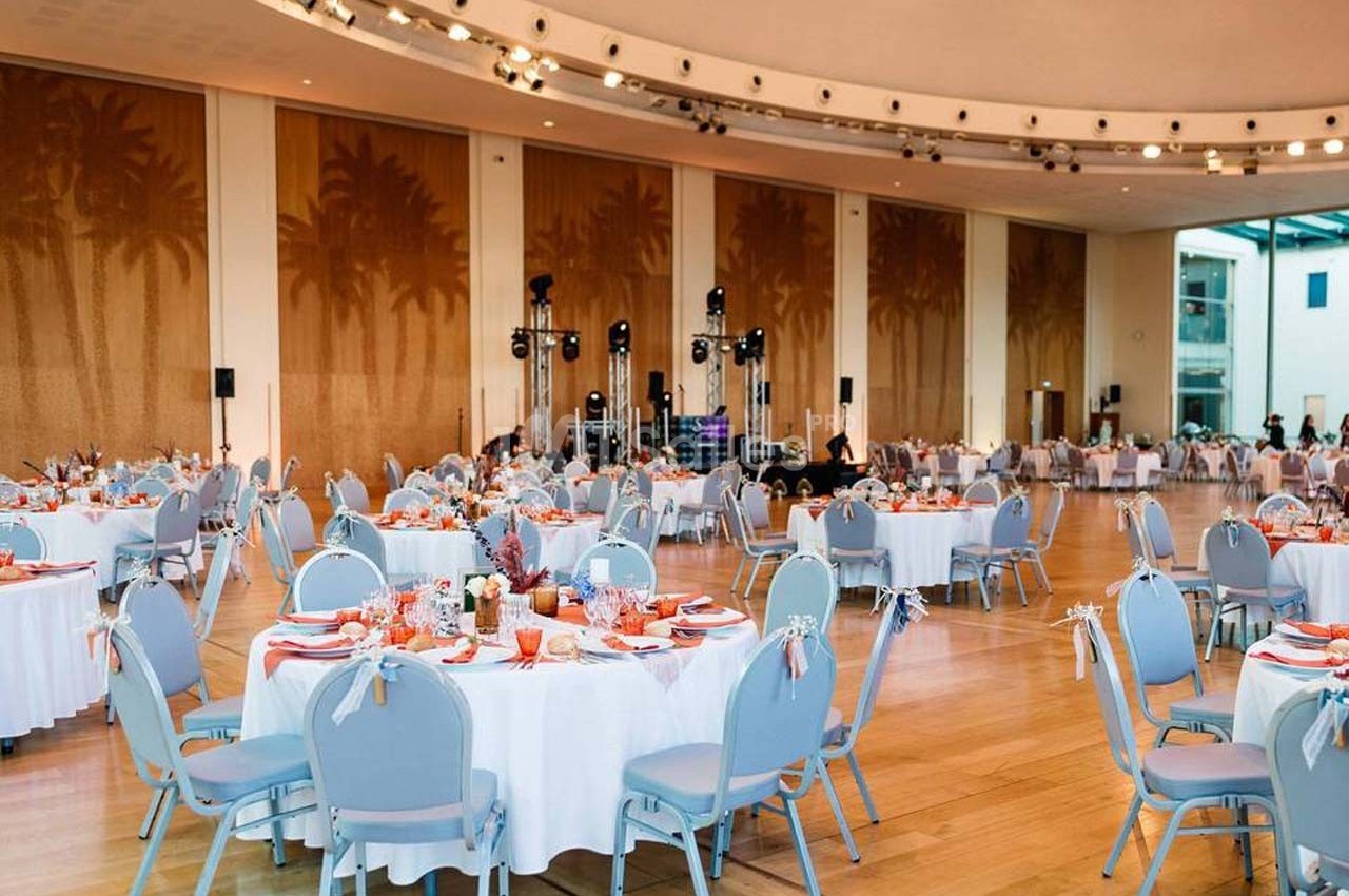 Salle de réception lumineuse avec tables rondes dressées, chaises bleues et décor mural représentant des palmiers.