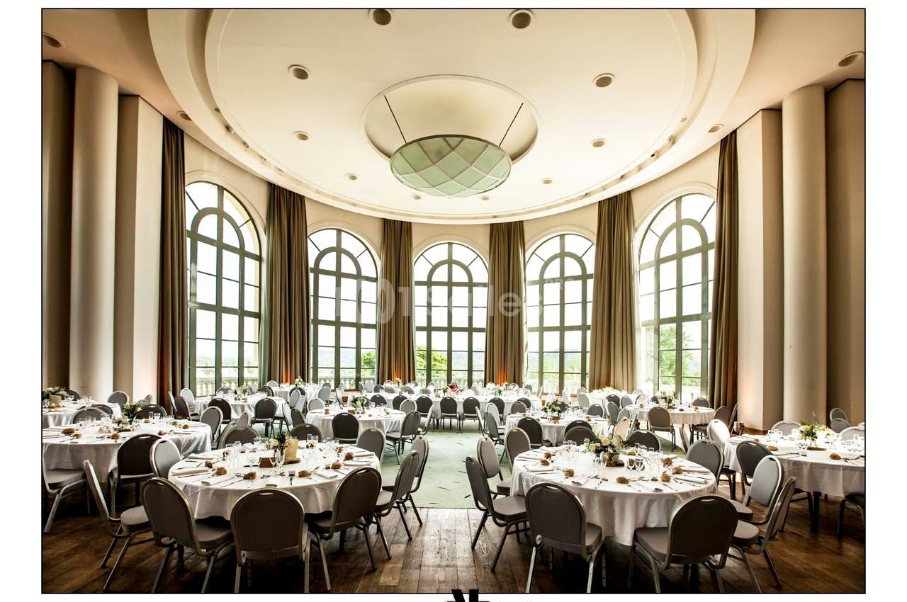 Salle de réception lumineuse avec de grandes fenêtres, des tables rondes dressées et des chaises grises.