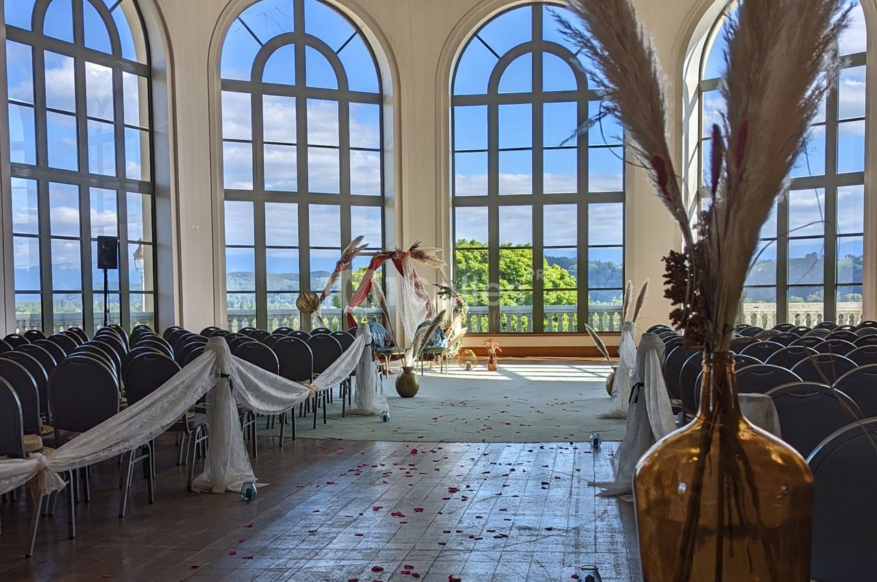 Salle de cérémonie lumineuse avec grandes fenêtres, rangées de chaises décorées et arche florale en arrière-plan.
