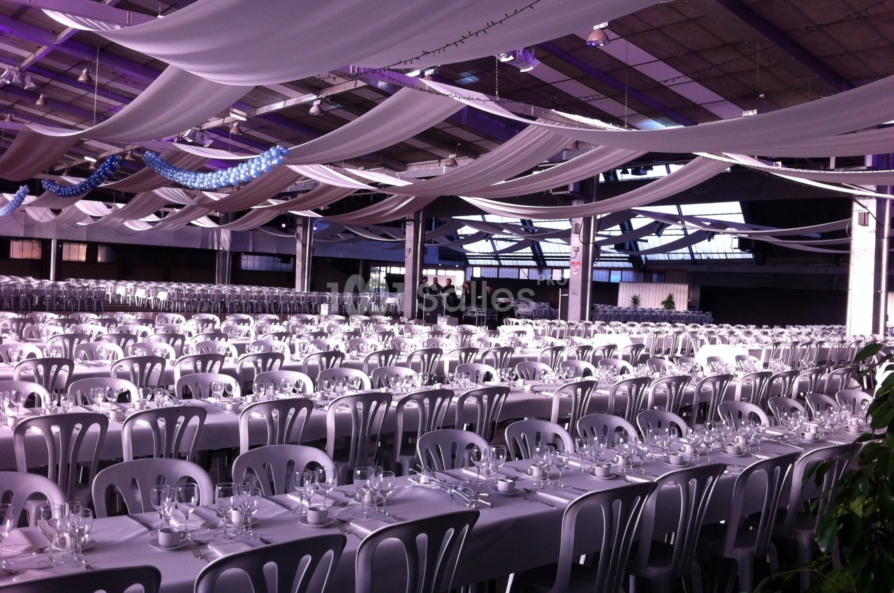 Grande salle décorée avec des drapés suspendus, aménagée avec de longues tables dressées et alignées pour un événement.