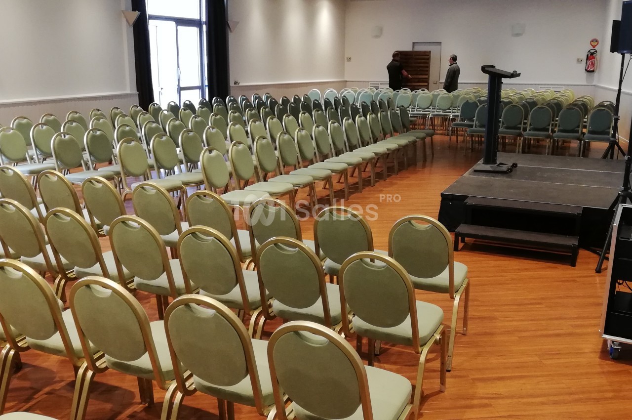 Salle de conférence avec rangées de chaises vertes alignées, une estrade avec pupitre et un équipement audio visible.