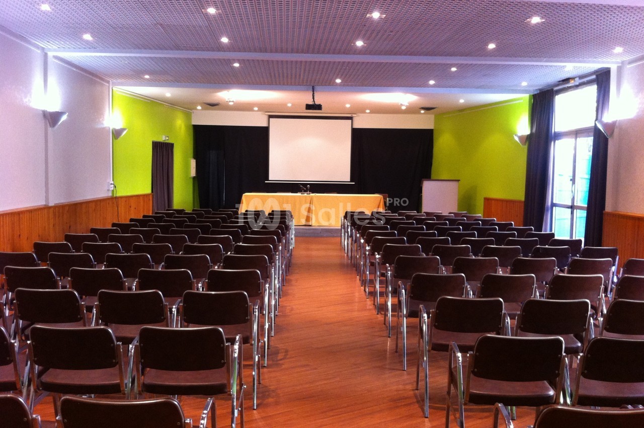 Salle de conférence avec rangées de chaises vides, écran de projection et table installée à l'avant.